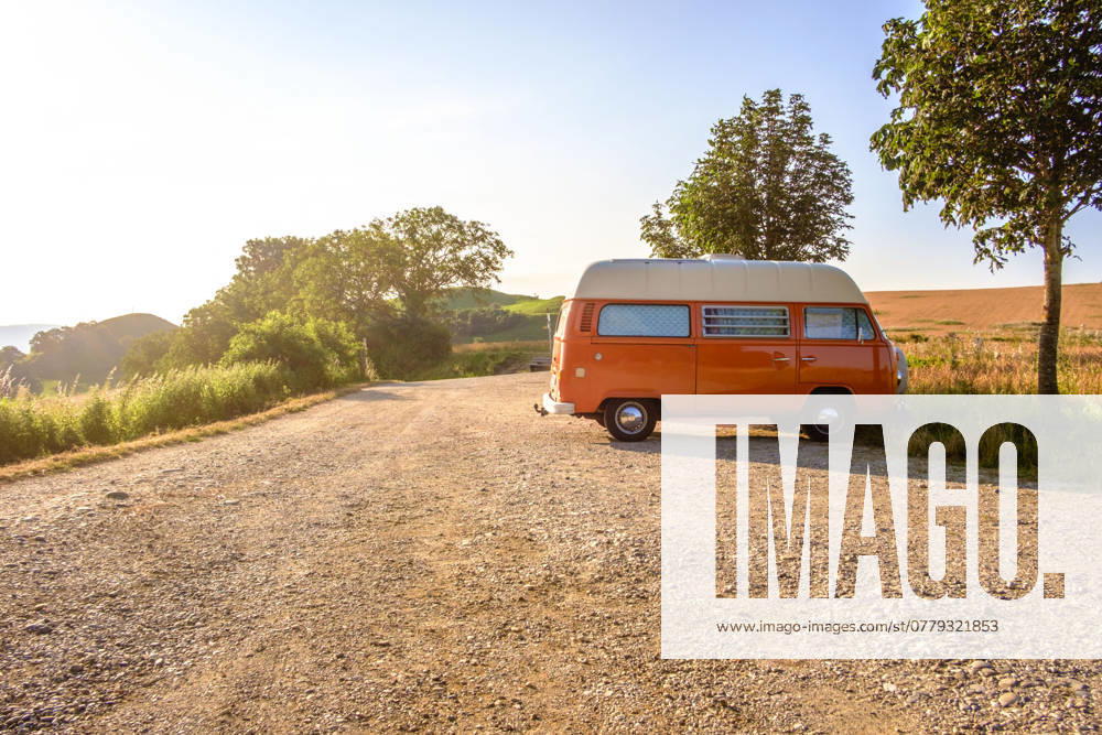 Ein Orange-weisser VW Bus Bulli steht auf einem Schotterparkplatz an einem Sommermorgen, Baden Württemberg, Deutschland. Ein Bulli steht in der Landschaft