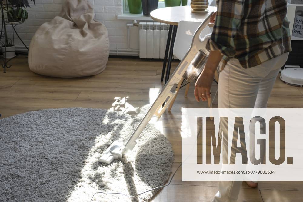 Eine Frau saugt einen runden Teppich in einem Haus zwischen Zimmerpflanzen mit einem Handstaubsauger. Allgemeine Reinigung des Hauses, Reinigungsservice und Hausfrau A woman vacuums a round carpet in a house among house plants with a hand vacuum cleaner. General cleaning of the house, cleaning service and housewife