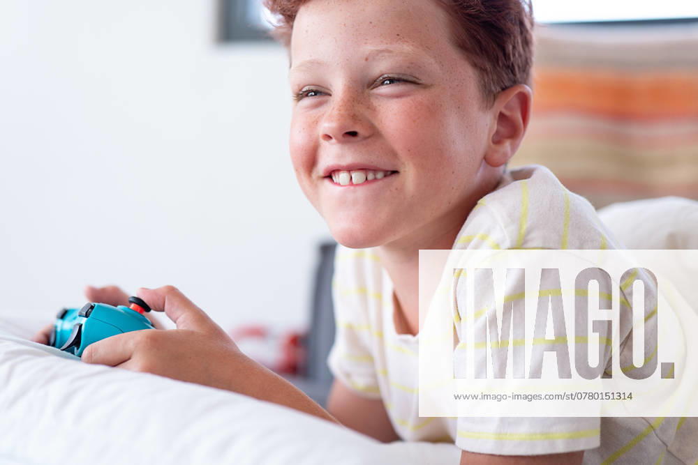 IMAGO Creative, Lifestyle, Gaming Young boy with red hair, visible freckles, and a big smile, playing video games on his handheld game console while lying on his stomach. He is wearing a light yellow striped t-shirt and looking slightly away from the camera. Model Released HodeiUnzueta_ID24741_716993_014