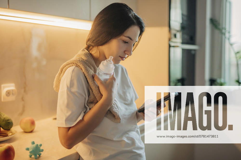 Young woman using smart phone and holding baby bottle in kitchen at home model released, Symbolfoto, VSNF02650