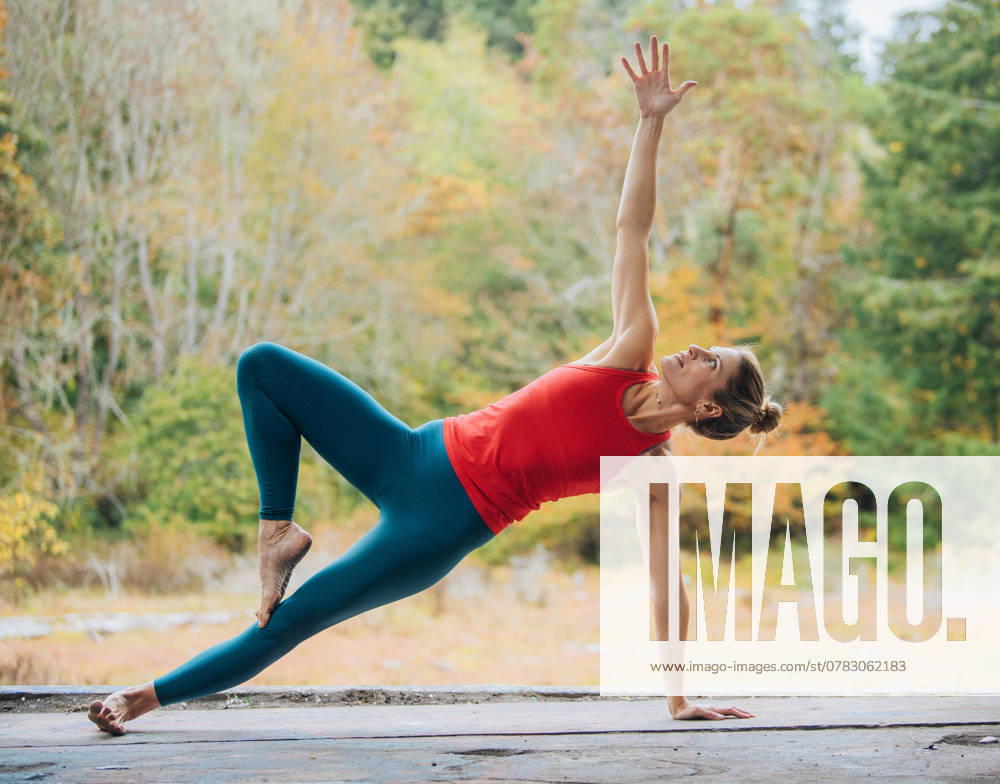 Woman performs a side plank yoga pose outdoors on a wooden deck with autumn trees in the background, USA sape00323