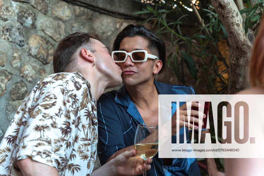 IMAGO Creative, Lifestyle, Feste im Garten #2 A tender scene captures one man kissing another on the cheek while holding glasses of wine at a lively garden party, reflecting a moment of love and celebration. Model Released DavidPrado_ID25521_737856_147