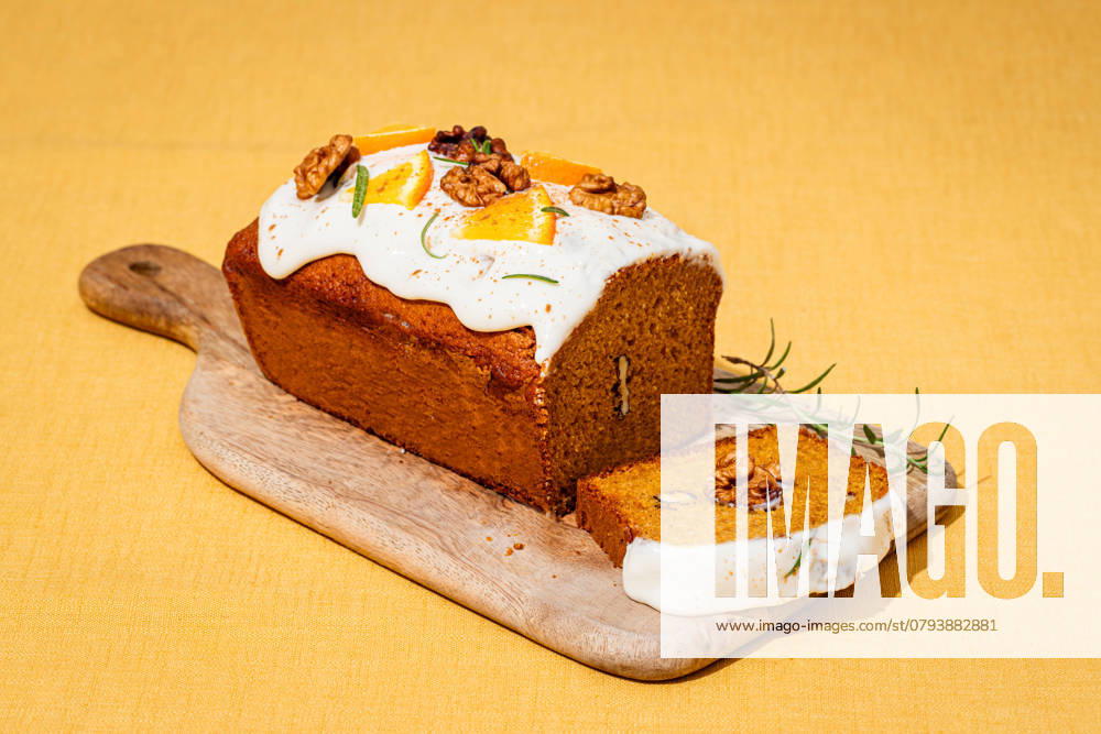 A freshly baked carrot cake loaf topped with cream cheese frosting, garnished with walnuts and rosemary Set on a wooden board against a yellow linen backdrop Model Released CintiaViudez_ID25764_744160_009