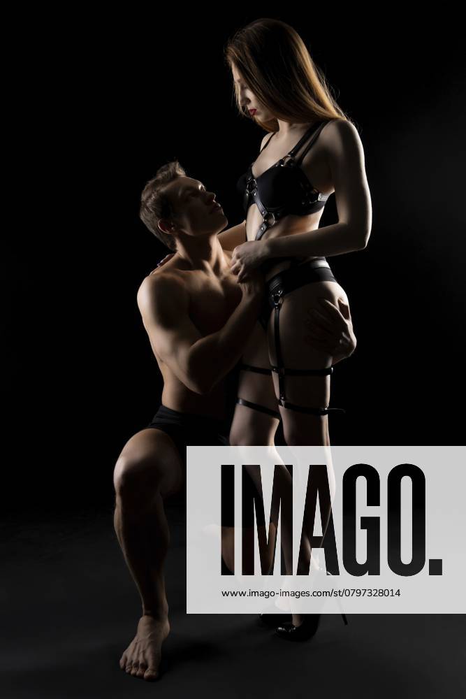 Seitenansicht einer jungen Frau in schwarzen Dessous und Ledergürteln, die einen Mann bei erotischen Spielen in einem dunklen Studio dominiert Side view of young female in black lingerie and leather belts dominating man during erotic games in dark studio