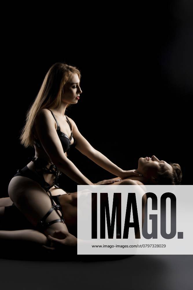 Seitenansicht einer jungen Frau in schwarzen Dessous, die einen Mann bei erotischen Spielen in einem dunklen Studio dominiert Side view of young female in black lingerie dominating man during erotic games in dark studio