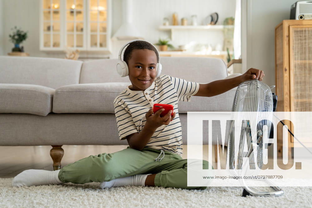 IMAGO Creative, Jahreszeit Sommer, Hitze Lächelnder, glücklicher afroamerikanischer Junge, der sich zu Hause vor einem elektrischen Ventilator entspannt Smiling happy African American boy child wearing headphones listening music or watching internet video while relaxing at home in front of electric fan. Children and summer heat ,model released, Symbolfoto