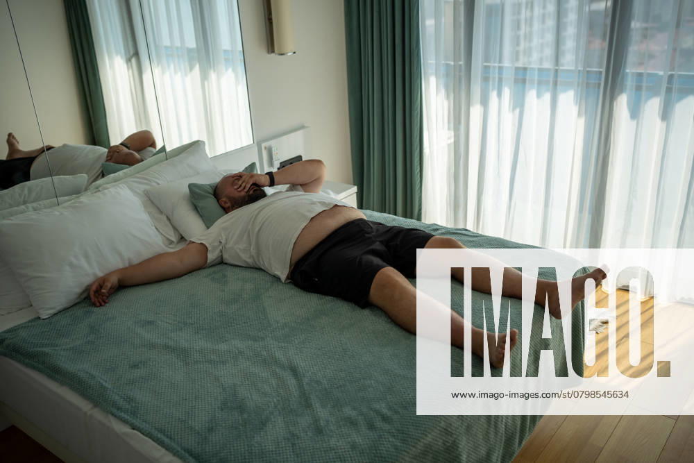 Ein fetter, übergewichtiger Mann stirbt vor Hitze. Faul, fettleibig übergewichtigen Mann liegt auf dem Bett, fühlt sich müde im Zimmer Fat man suffers dying from heat. Lazy, obese overweight young guy lies on bed, suffering from summer abnormal warmth, feels tired, unpleasant discomfort sensations in body, high blood pressure. ,model released, Symbolfoto