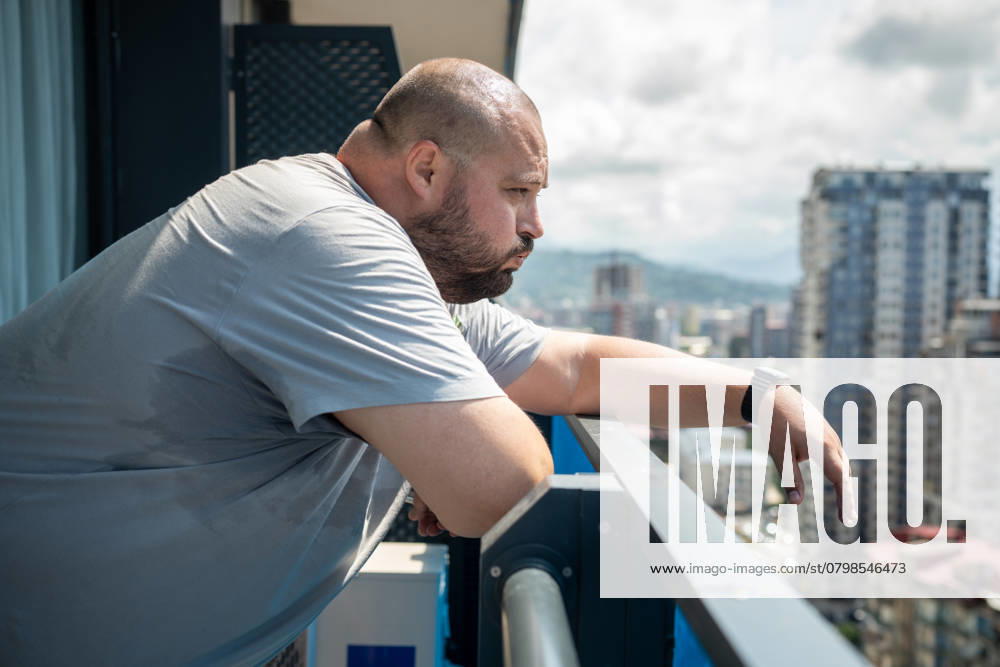 IMAGO Creative, Jahreszeit Sommer, Hitze Übergewichtiger Mann, der sich bei sommerlicher Hitze schlecht fühlt, steht auf einem Balkon und schaut auf die Straße. Overweight man feeling bad from heat in summer standing on balcony looking at street in summer. High sweating, hyperhidrosis, obesity, heaviness from hot weather high humidity, excess body weight. ,model released, Symbolfoto