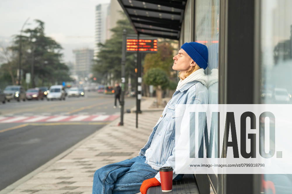 IMAGO Creative, Gesellschaft, Leisure Sickness Müder Mann mit geschlossenen Augen wartet auf den Transport an der Bushaltestelle am Morgen nach der Nachtarbeit. Tired sleepy young man sitting at bus stop with cup coffee, relaxing in warm sunlight with eyes closed. Early morning of exhausted student guy after sleepless night waiting for transport to college. ,model released, Symbolfoto