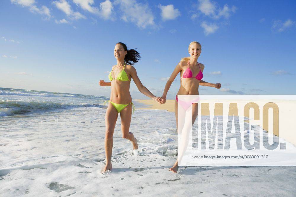 Two women dressed in bikinis jogging along the coastal line and holding their hands A5