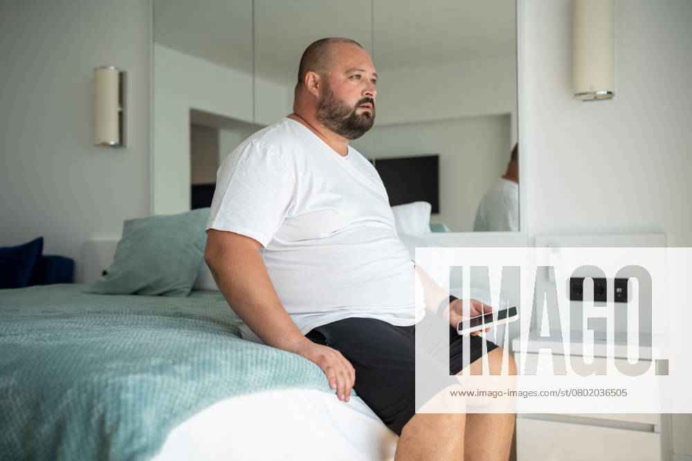 IMAGO Creative, Gesellschaft, Leisure Sickness Ein fauler, übergewichtiger Mann sitzt mit dem Telefon auf einem Hotelbett und blickt nachdenklich auf eine Stelle. Einsamkeit, Angst Ein fauler, übergewichtiger Mann sitzt mit dem Telefon auf einem Hotelbett und blickt nachdenklich auf eine Stelle. Einsamkeit, Angst ,model released, Symbolfoto