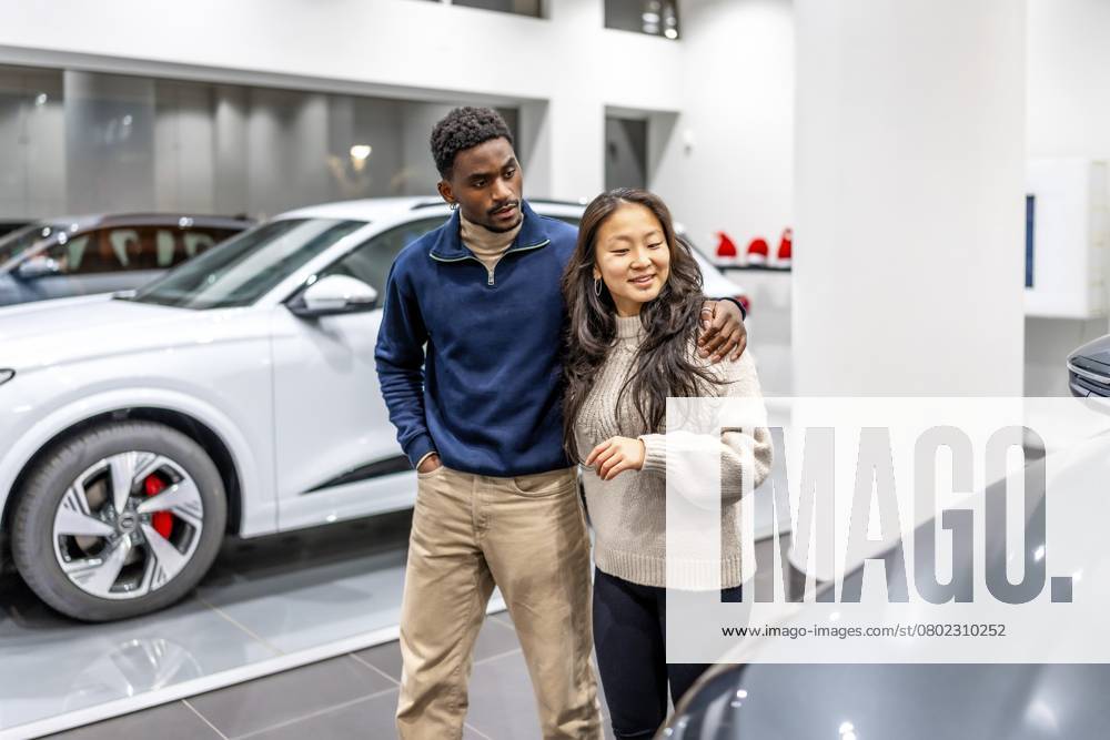 Ein glückliches multiethnisches Paar, das im Autohaus ein neues Elektroauto kauft, umarmt sich und wählt sein zukünftiges Fahrzeug Happy multiethnic couple buying a new electric car at the dealership, embracing and choosing their future vehicle
