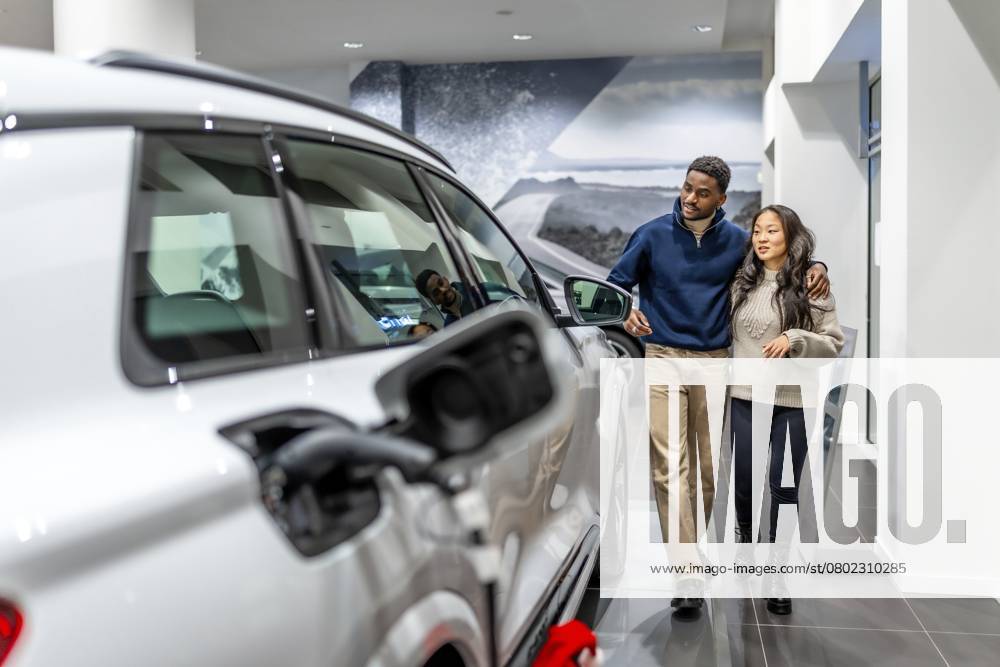 Ein multiethnisches Paar geht durch ein Autohaus und wählt fröhlich sein neues Elektrofahrzeug aus, während es den modernen Ausstellungsraum genießt Multiethnic couple walking through a car dealership, happily selecting their new electric vehicle while enjoying the modern showroom experience