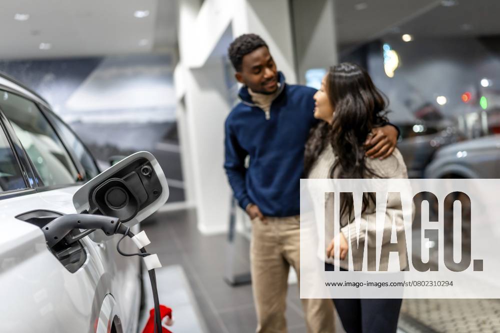 Ein multiethnisches Paar umarmt sich bei der Auswahl eines neuen Elektroautos in einem Autohaus, das sich auf nachhaltige Mobilität konzentriert Multiethnic couple embracing while choosing a new electric car in a dealership, focusing on sustainable transportation