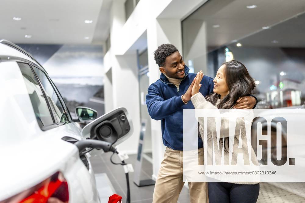 Ein glückliches multiethnisches Paar feiert den Kauf seines neuen Elektroautos mit einem High Five im Autohaus und strahlt vor Freude und Begeisterung Happy multiethnic couple celebrating their new electric car purchase with a high five at the dealership, radiating joy and excitement