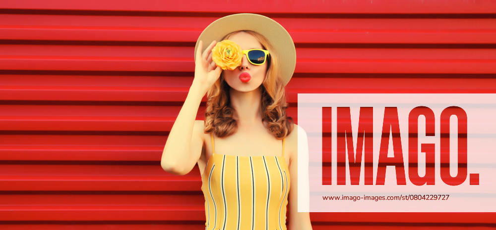 Sommer Porträt der stilvollen jungen Frau bläst Kuss mit gelben Blütenknospen auf rotem Hintergrund Summer portrait of stylish young woman blowing kiss with yellow flower buds looking for something in round straw hat on red background ,model released, Symbolfoto