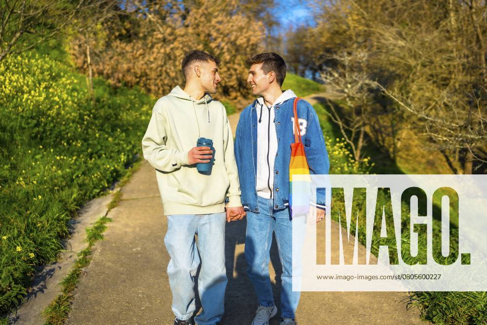 Junges weißes schwules Paar, das sich an den Händen hält und in einem Park spazieren geht, um einen sonnigen Tag zu genießen Young caucasian gay couple holding hands and walking in a park, enjoying a sunny day together