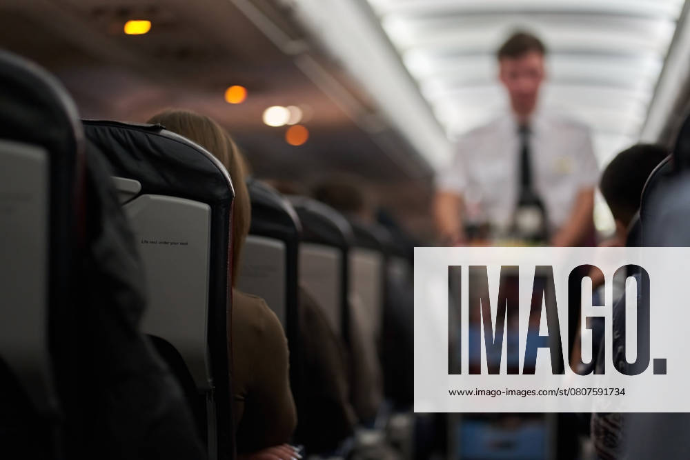 A male steward who distributes food and drinks on board the aircraft. The passengers are sitting in their seats and watching. View along the row A male steward who distributes food and drinks on board the aircraft. The passengers are sitting in their seats and watching. View along the row