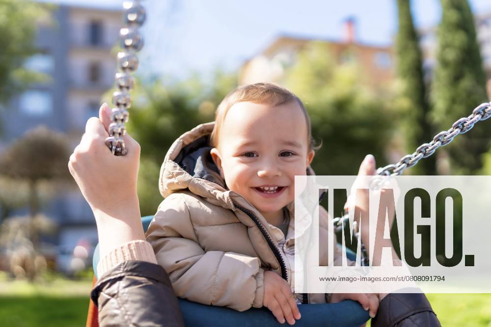 Lächelnder kleiner Junge, der von seiner Mutter auf dem Spielplatz an einem sonnigen Tag auf einer Schaukel geschoben wird Smiling baby boy enjoying a swing ride pushed by his mother at the playground on a sunny day