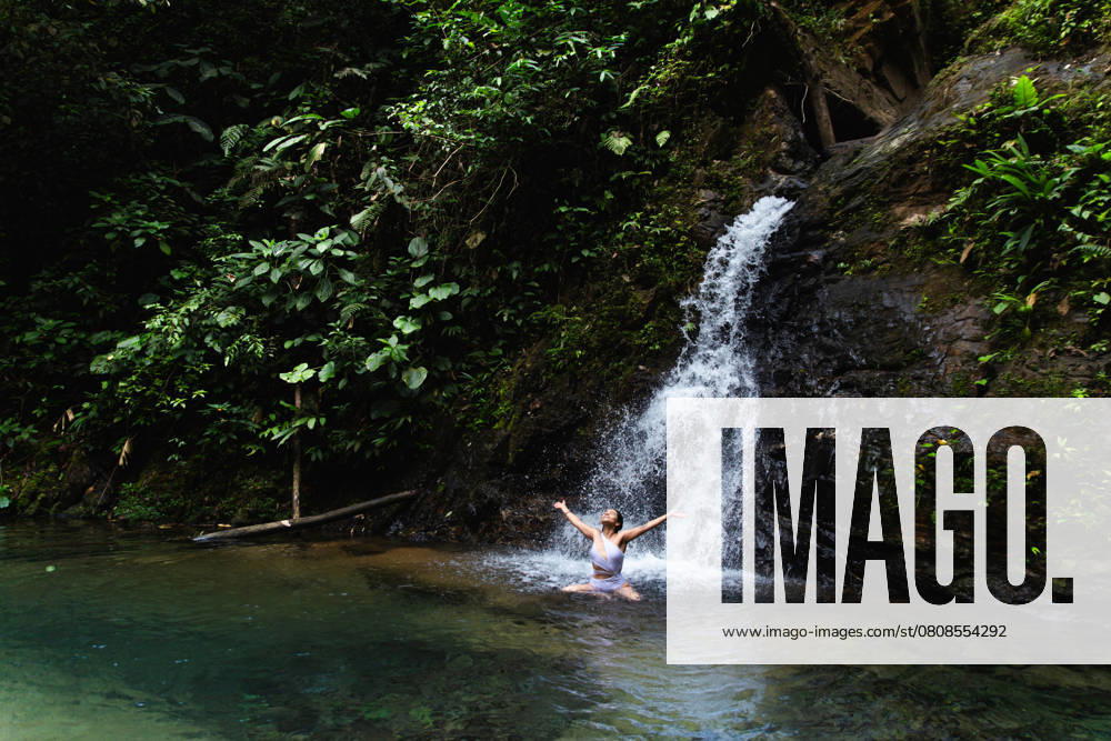 Young woman enjoying and stretching arms in a natural pool under a waterfall in the Choco Andino rainforest, Ecuador. This image captures the spirit of ecotourism and adventure in a biodiverse tropical landscape. Model Released Cardellach_ID27637_793647_017