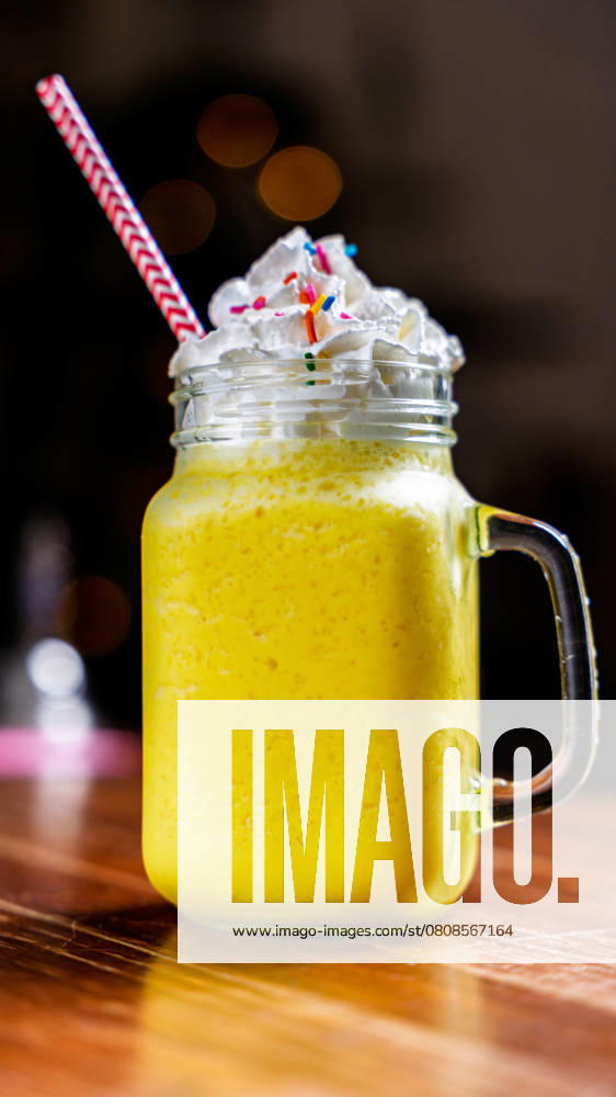 A vibrant mango smoothie topped with creamy whipped cream and colorful sprinkles, served in a mason jar with a striped straw on a wooden cafe table. Perfect for summer vibes. Model Released LeoTenorio_ID27358_784894_019