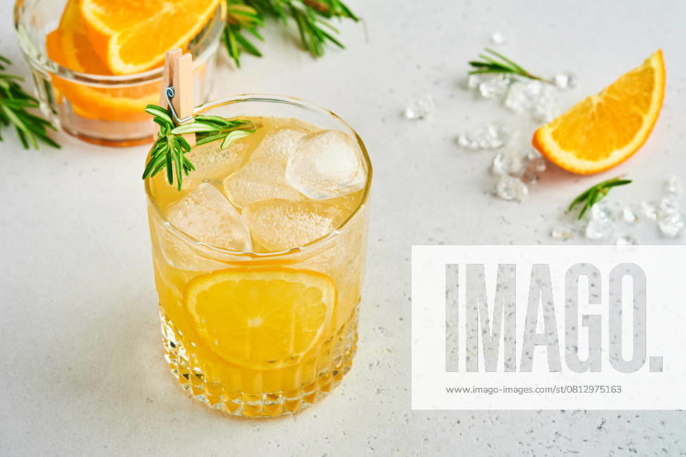 Orangensaft oder Cocktail mit Rosmarin und Orange mit Eis im Glas, kalte Sommerlimonade auf hellgrauem Schiefer, Stein oder Beton Hintergrund. Strandkonzept mit tiefem Sonnenschatten. Ansicht von oben. Orange juice or cocktail with rosemary and orange with ice in glass, cold summer lemonade on light gray slate, stone or concrete background. Beach concept with deep sun shadow. Top view. (License=RF)