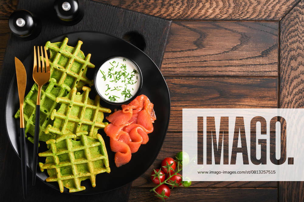 Grüne belgische Waffeln. Spinat-, Bärlauch- oder Pesto-Waffeln mit rotem Lachs und Sahnesauce auf schwarzem Teller vor dunklem, rustikalem Holzhintergrund. Leckeres Frühstück, Snack, Brunch. Ansicht von oben. Attrappe Green Belgian waffles. Spinach or wild garlic or pesto waffles with red salmon and cream sauce on black plate over dark rustic wooden background. Delicious breakfast, snack, brunch. Top view. Mock up (License=RF)