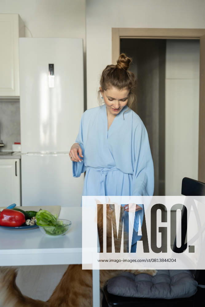 MR_ID27929_20617_TatyanaRangulova.jpg, PR_ID27929_10891_Cat.jpg, PR_ID27929_10892_Room.jpg A woman in a light blue robe feeds her cat in a modern kitchen. Fresh vegetables on the table create a warm, homely atmosphere, perfect for a lifestyle theme., Property released Model Released LiliyaSolopova_ID27929_802056_002