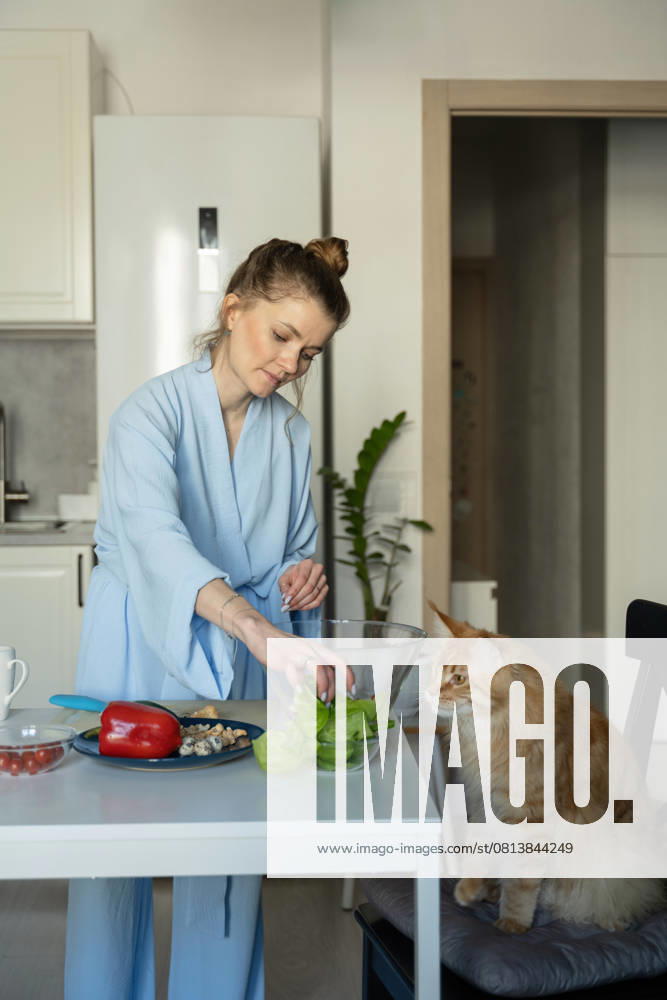 MR_ID27929_20617_TatyanaRangulova.jpg, PR_ID27929_10891_Cat.jpg, PR_ID27929_10892_Room.jpg A woman in a blue robe prepares vegetables at a kitchen table. A curious cat sits nearby, observing her actions. The scene captures a cozy, domestic moment., Property released Model Released LiliyaSolopova_ID27929_802060_006