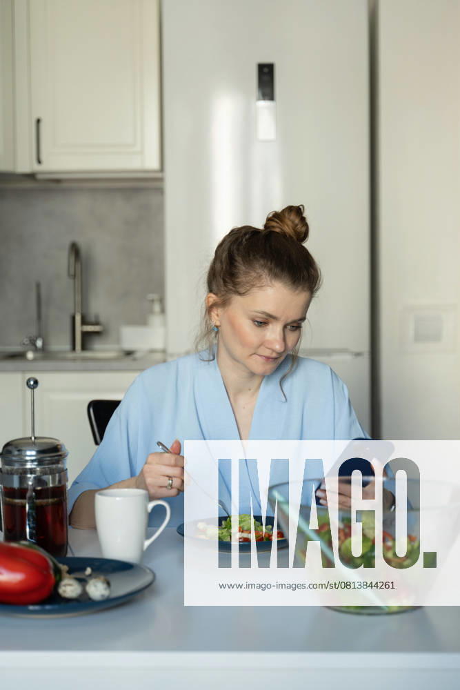 MR_ID27929_20617_TatyanaRangulova.jpg, PR_ID27929_10891_Cat.jpg, PR_ID27929_10892_Room.jpg A woman wearing a blue robe is sitting at a table in her kitchen, eating a fresh salad while looking at her smartphone. The scene conveys a relaxed domestic atmosphere., Property released Model Released LiliyaSolopova_ID27929_802062_007