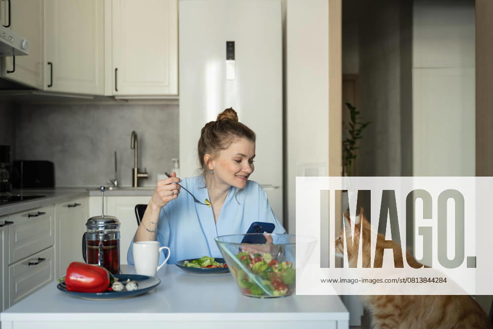 MR_ID27929_20617_TatyanaRangulova.jpg, PR_ID27929_10891_Cat.jpg, PR_ID27929_10892_Room.jpg A woman sits at a kitchen table, enjoying a fresh salad while checking her phone. A curious cat sits nearby, capturing the cozy atmosphere of home life., Property released Model Released LiliyaSolopova_ID27929_802065_009