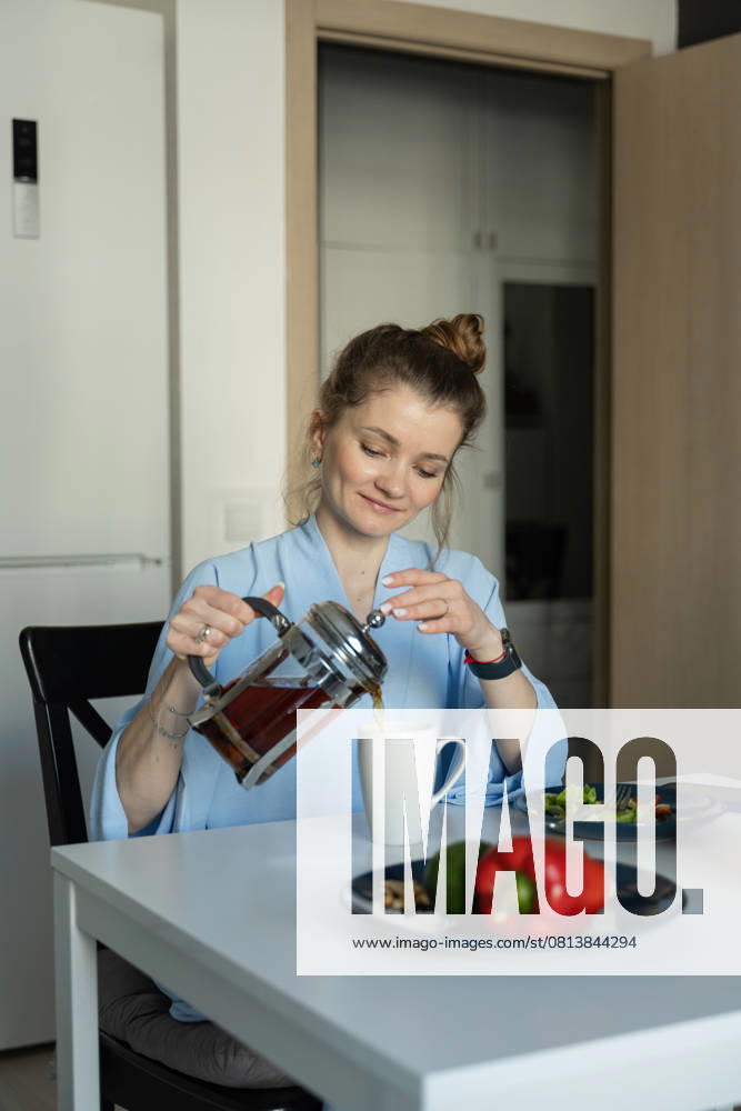 MR_ID27929_20617_TatyanaRangulova.jpg, PR_ID27929_10891_Cat.jpg, PR_ID27929_10892_Room.jpg A woman in a casual setting pours tea into a mug at her kitchen table, creating a warm and cozy atmosphere. Fresh vegetables are seen on the table, enhancing the homely vibe., Property released Model Released LiliyaSolopova_ID27929_802066_010