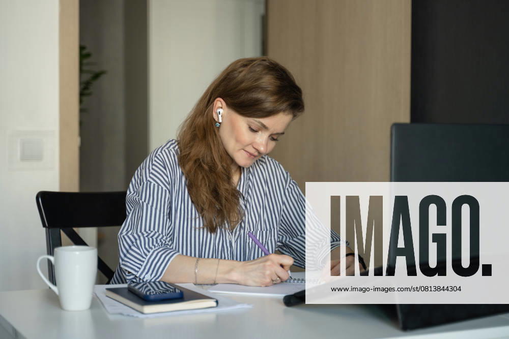 MR_ID27929_20617_TatyanaRangulova.jpg, PR_ID27929_10891_Cat.jpg, PR_ID27929_10892_Room.jpg A woman sits at a desk at home, writing in a notebook. She is wearing wireless earphones, a striped shirt, and is focused on her laptop. A mug and mobile phone are nearby., Property released Model Released LiliyaSolopova_ID27929_802068_011