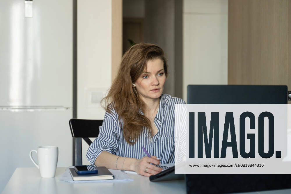 MR_ID27929_20617_TatyanaRangulova.jpg, PR_ID27929_10891_Cat.jpg, PR_ID27929_10892_Room.jpg Woman in striped shirt works at home, focused on laptop. She writes in a notebook with a cup on the table, symbolizing work-from-home productivity., Property released Model Released LiliyaSolopova_ID27929_802069_012