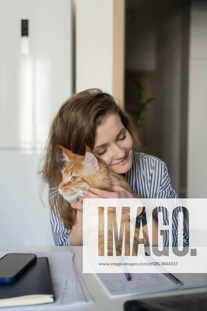 MR_ID27929_20617_TatyanaRangulova.jpg, PR_ID27929_10891_Cat.jpg, PR_ID27929_10892_Room.jpg A woman in a striped shirt lovingly holds her ginger cat while working, surrounded by a phone and a notebook in a bright home setting, conveying warmth and relaxation., Property released Model Released LiliyaSolopova_ID27929_802072_014