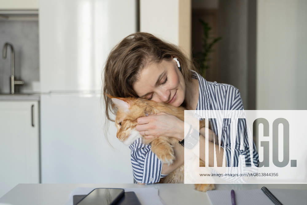 MR_ID27929_20617_TatyanaRangulova.jpg, PR_ID27929_10891_Cat.jpg, PR_ID27929_10892_Room.jpg A woman embraces a ginger cat while sitting at a table in a cozy kitchen. She wears striped clothing and earbuds, exuding warmth and contentment in a relaxing home environment., Property released Model Released LiliyaSolopova_ID27929_802076_016