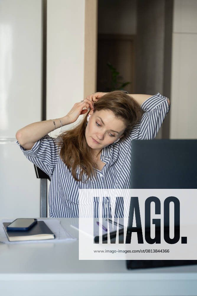 MR_ID27929_20617_TatyanaRangulova.jpg, PR_ID27929_10891_Cat.jpg, PR_ID27929_10892_Room.jpg Woman in a striped shirt at home, adjusting her earphones while working on a laptop. A phone, notebook, and pen are on the table, creating a cozy workspace., Property released Model Released LiliyaSolopova_ID27929_802077_017