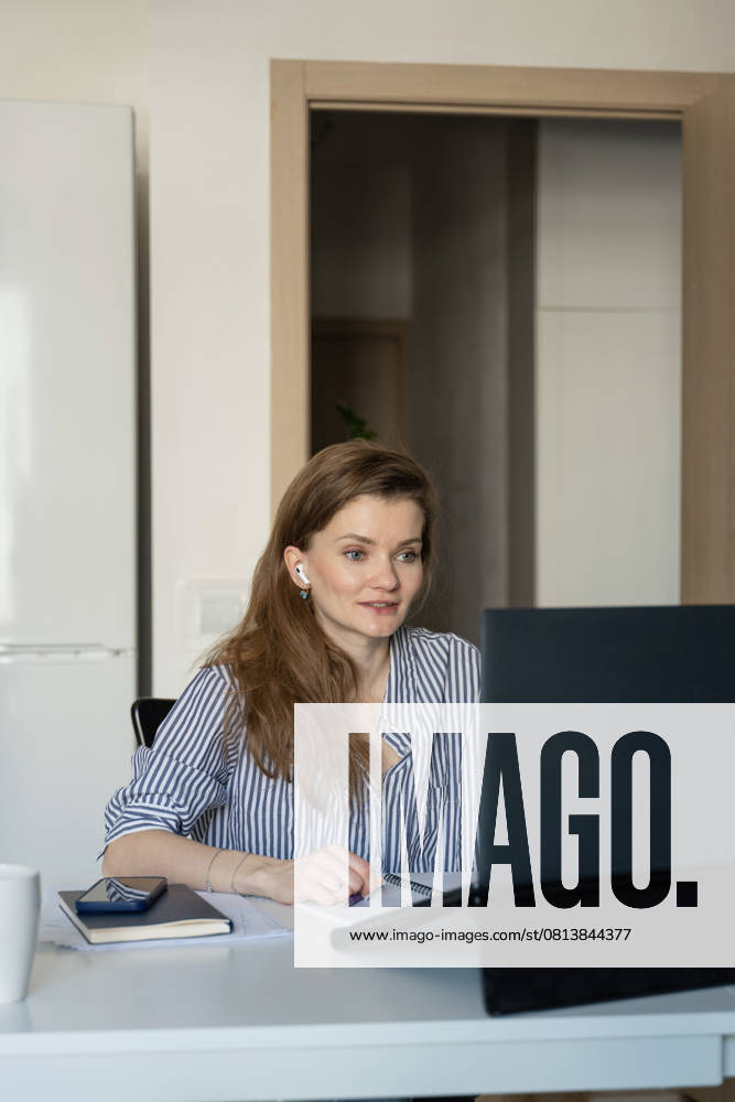 MR_ID27929_20617_TatyanaRangulova.jpg, PR_ID27929_10891_Cat.jpg, PR_ID27929_10892_Room.jpg A woman in a striped shirt works on a laptop at home, engaging in a virtual meeting. She appears focused and attentive, using earbuds and taking notes with a notebook nearby., Property released Model Released LiliyaSolopova_ID27929_802078_018