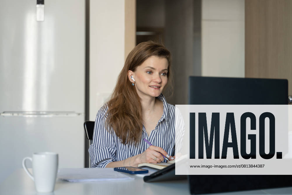 MR_ID27929_20617_TatyanaRangulova.jpg, PR_ID27929_10891_Cat.jpg, PR_ID27929_10892_Room.jpg A woman sits at a table working remotely from home. She writes in a notebook while focusing on her laptop screen, with a mug and phone nearby., Property released Model Released LiliyaSolopova_ID27929_802080_019