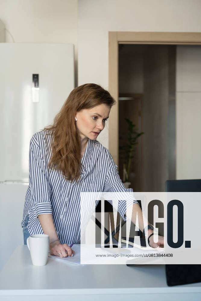 MR_ID27929_20617_TatyanaRangulova.jpg, PR_ID27929_10891_Cat.jpg, PR_ID27929_10892_Room.jpg A woman dressed casually in stripes works from home, standing at a table with a laptop, papers, and a phone, illustrating modern remote work and concentration., Property released Model Released LiliyaSolopova_ID27929_802082_021