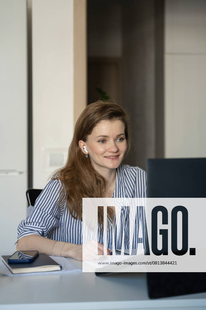 MR_ID27929_20617_TatyanaRangulova.jpg, PR_ID27929_10891_Cat.jpg, PR_ID27929_10892_Room.jpg A woman sitting at her desk at home, smiling as she looks at her laptop screen. She is wearing earbuds, suggesting remote communication or entertainment., Property released Model Released LiliyaSolopova_ID27929_802083_022