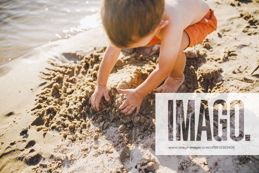Ein kleiner Junge baut bei Sonnenuntergang am Ufer des Teiches Figuren aus dem Sand, die Hände graben den Sand in einem knackigen Plan um A little boy builds figures from the sand on the shore of the pond at sunset of the day, hands dig up the sand in a crisp plan