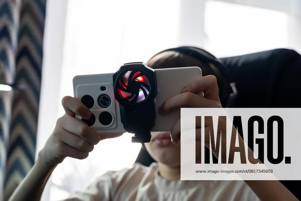 IMAGO Creative, Lifestyle, Gaming A teen boy plays on a smartphone equipped with an external cooling fan attachment while wearing headphones. The setup reflects immersive mobile gaming in a tech-savvy, indoor environment Model Released Ikvyatkovskaya_ID28180_830161_028