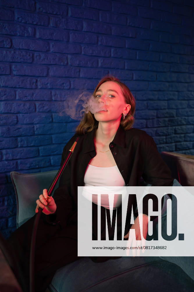 PR_ID27963_10908_Cafe.jpg, MR_ID27963_20660_AlenaZhdanova.jpg A relaxed young woman in a stylish outfit enjoys smoking hookah in a trendy lounge. She exhales smoke against a vibrant blue brick wall, creating a calm atmosphere., Property released Model Released LiliyaSolopova_ID27963_803186_007