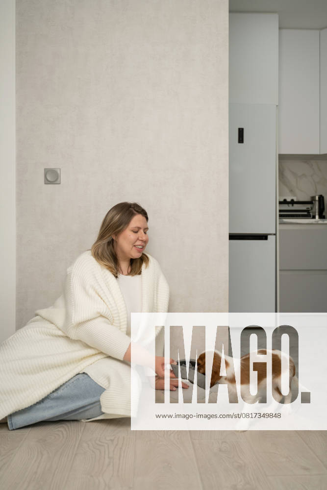 A woman in a knitted cardigan sits on the floor in a modern kitchen, feeding her puppy. The scene captures a warm, homely atmosphere, emphasizing a bond with pets Model Released LiliyaSolopova_ID28310_834495_037