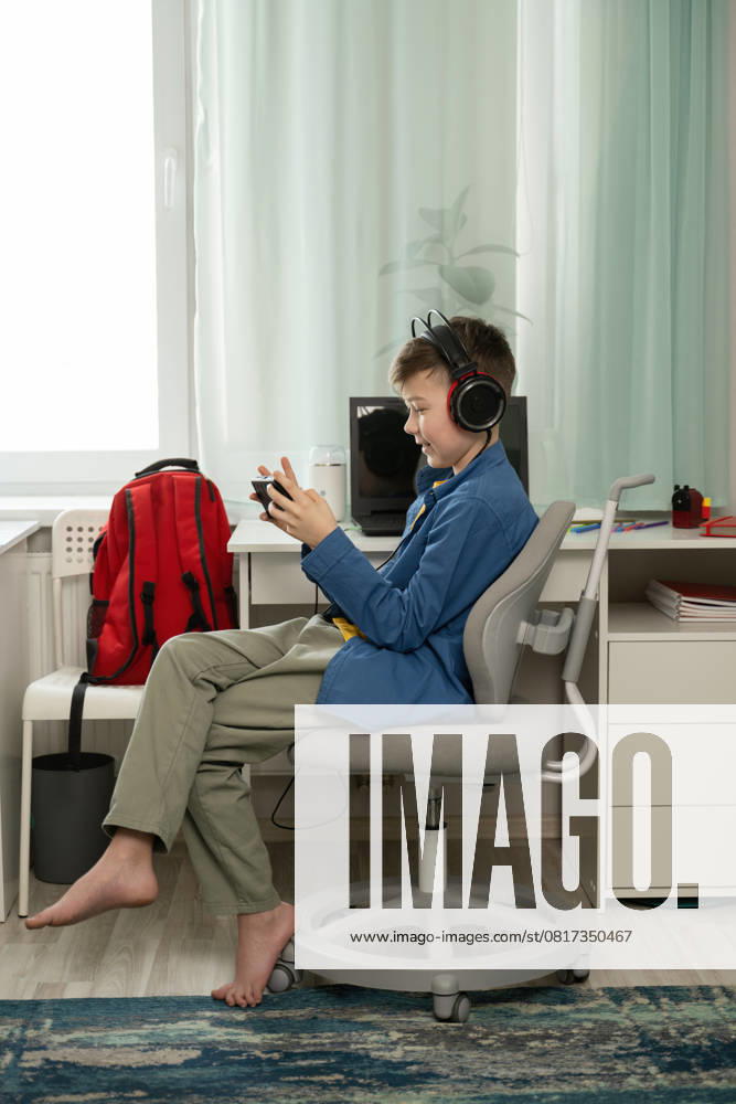 IMAGO Creative, Lifestyle, Gaming MR_ID28313_20909_IgorSushin.jpg, PR_ID28313_10984_Room.jpg, MR_ID28313_20910_AnnaSushina.jpg A young boy sits barefoot, focused on his mobile game. Wearing headphones, he enjoys gaming in his comfortable room with a backpack nearby and a tidy desk, Property released Model Released LiliyaSolopova_ID28313_834569_036
