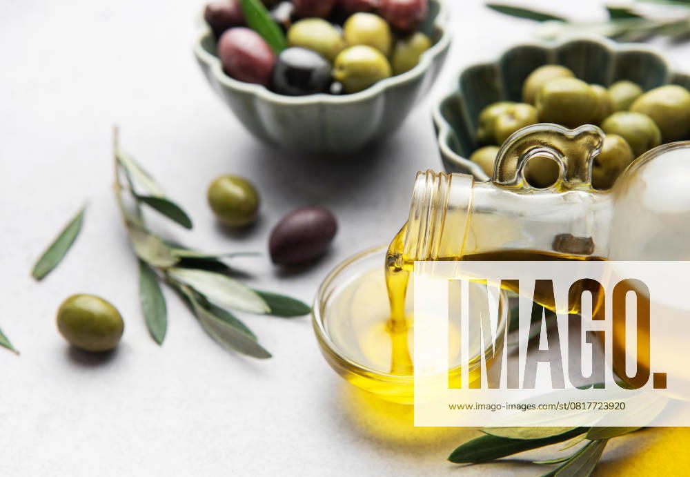 Natives Olivenöl in eine kleine Schale mit Oliven und Olivenzweigen gießen Golden olive oil pouring from a glass bottle into a small bowl, surrounded by fresh olives and olive branches (License=RF)