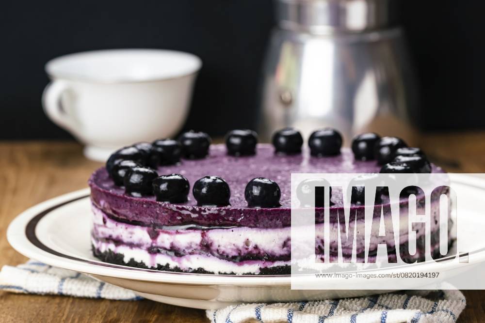 Seitenansicht eines köstlichen hausgemachten Blaubeer-Käsekuchens, garniert mit eingelegten Blaubeeren, in einer braunen Keramikschale mit Mokakanne und weißer Keramiktasse auf einem Holztisch Side view of delicious homemade blueberry cheesecake garnished with preserved blueberry in brown ceramic dish with moka pot coffee maker and white ceramic coffee cup on wooden table