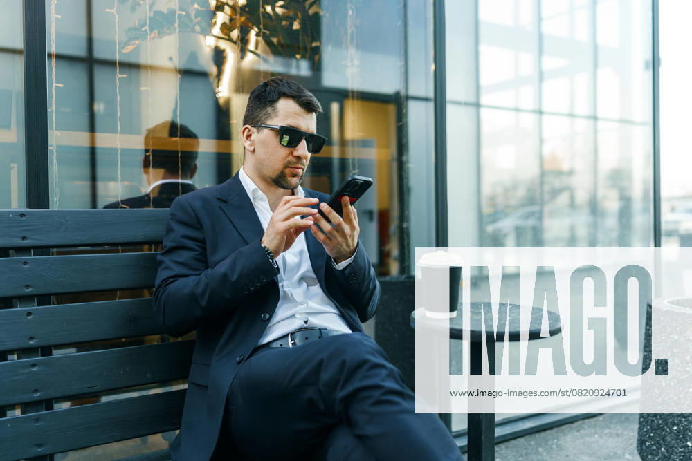 A businessman in a suit sits outdoors, using a smartphone while enjoying coffee. The modern urban setting highlights a blend of work and leisure. Sunglasses add sophistication Model Released PavelG_ID28721_848567_012