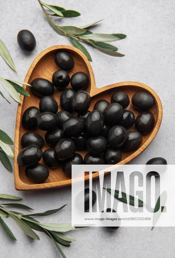 Fresh black olives in heart shaped wooden bowl on gray background with olive branches and copy space Model Released OlenaRudo_ID28871_858125_066
